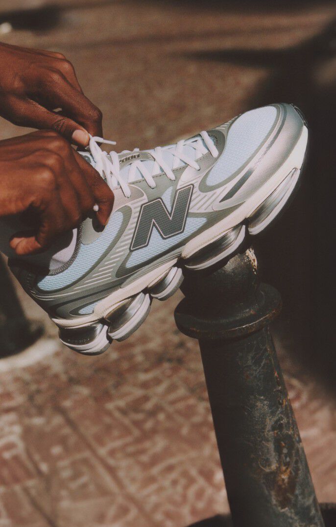 A close up of a person tying their ABZORB 2000 New Balance shoes in light chrome blue and silver metallic with a blurry city backgound 
