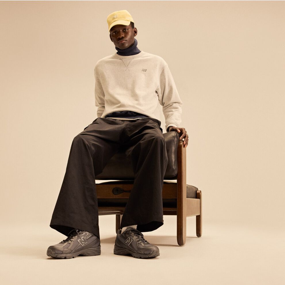 Model sitting on wooden and leather chair behind a beige background wearing a tan shirt, yellow hat, and dark New Balance 740s. 