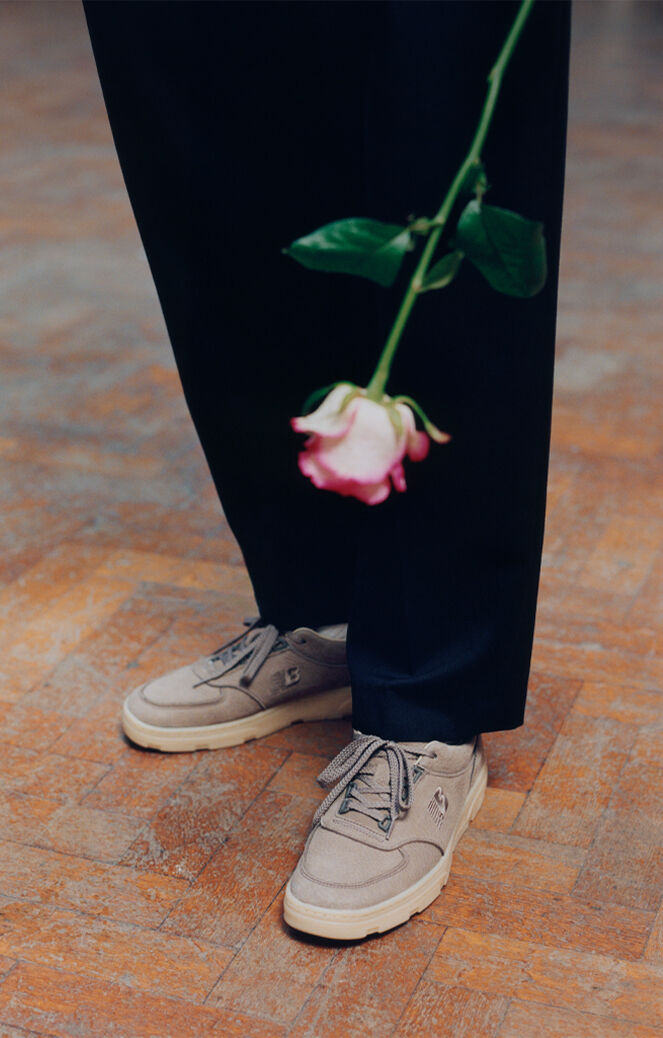Knee down view of a man wearing a pair of New Balance Made in UK sneakers standing on a herringbone pattern floor with a white and pink rose being held in frame.