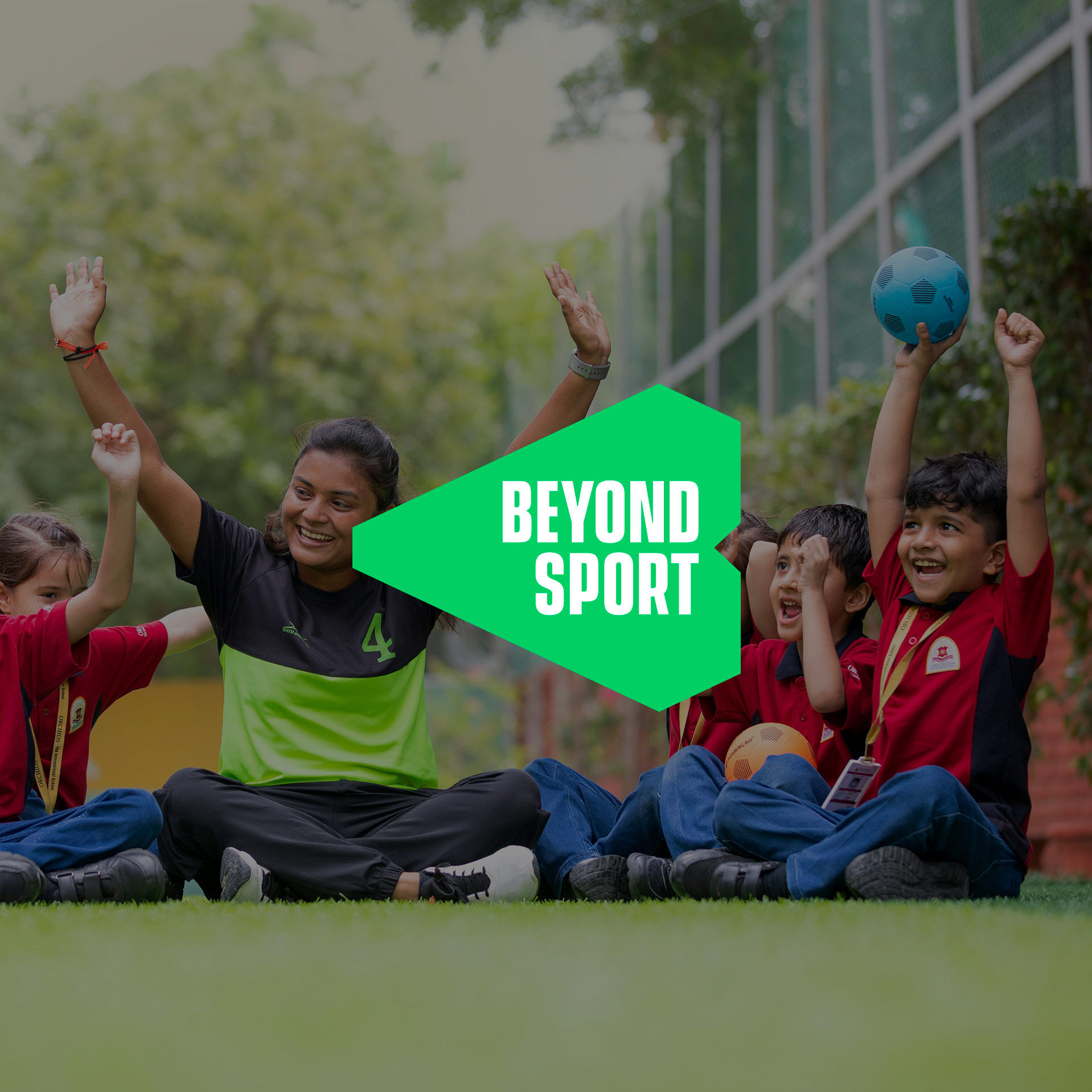 A group of medium-skinned children sitting in a semi-circle with a female-presenting adult learning about soccer and cheering, with arms happily raised to the sky. 