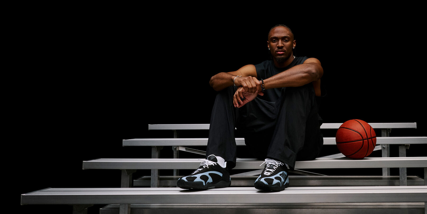 Person seated on metal bleachers beside a basketball, highlighting black-and-light-blue athletic sneakers in a dark studio setting. 