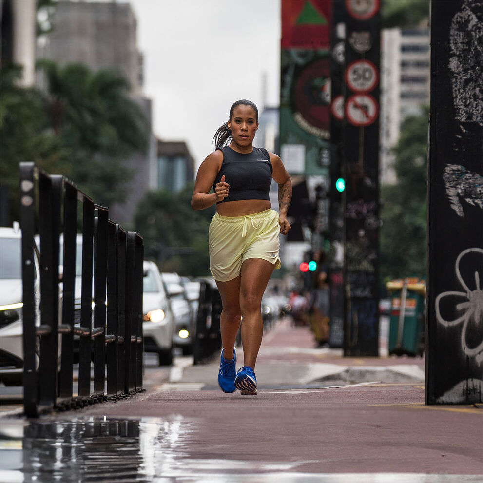 A woman running towards the camera through a cityscape, wearing New Balance running apparel, with tall buildings and urban streets in the background.