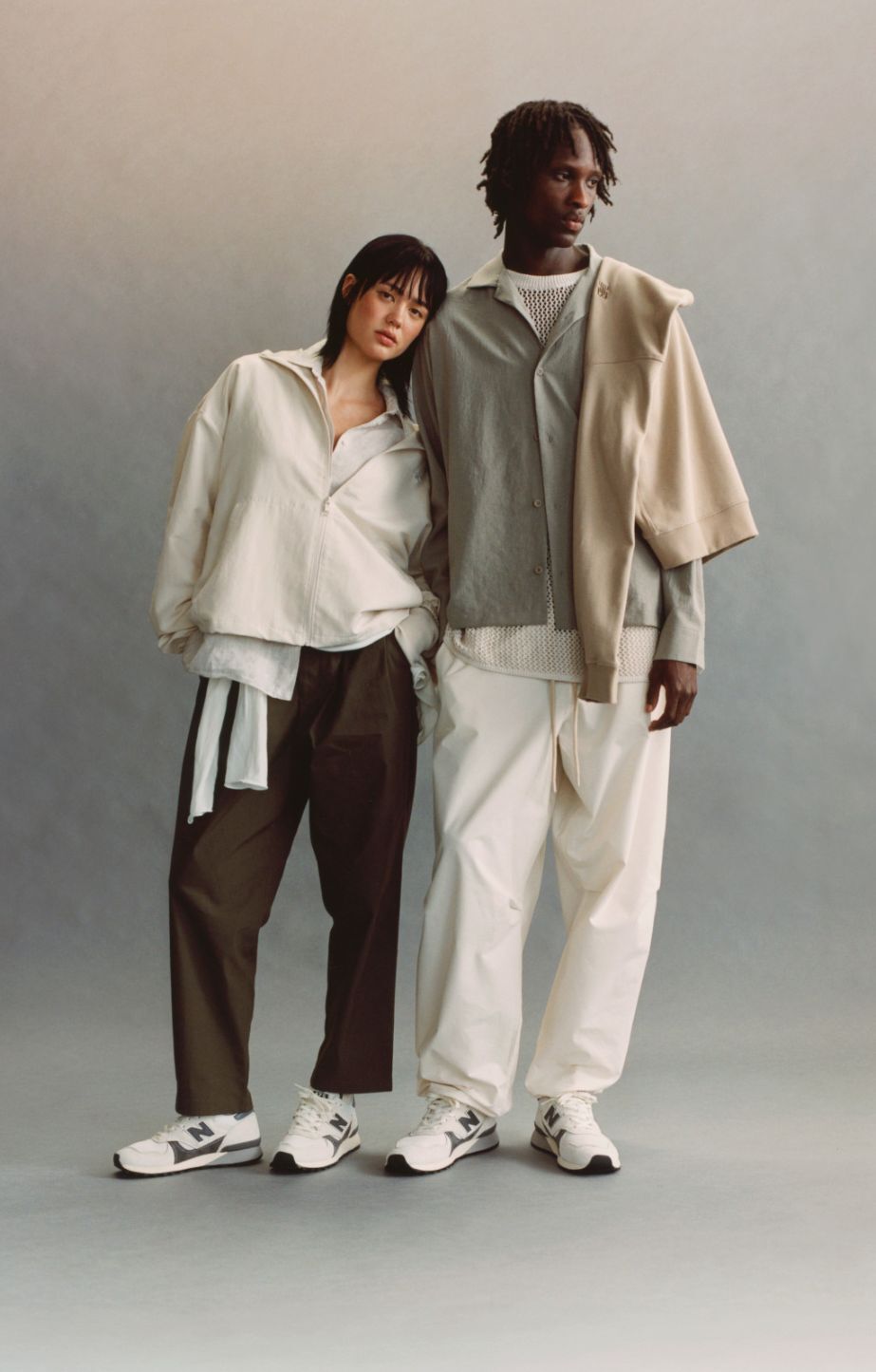 A man and a woman standing on a grey studio background, both wearing New Balance 475 shoes.