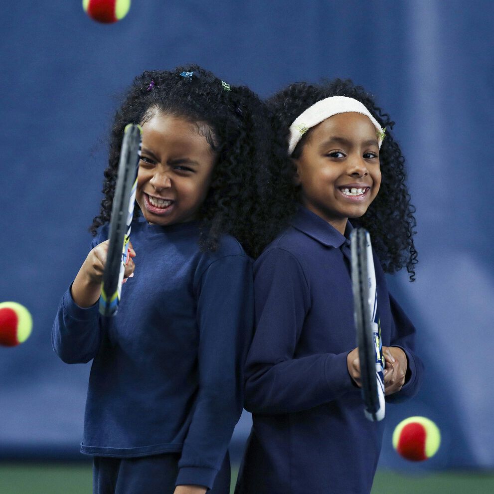 Two dark - skinned young female - presenting child holding tennis racquets and smiling.