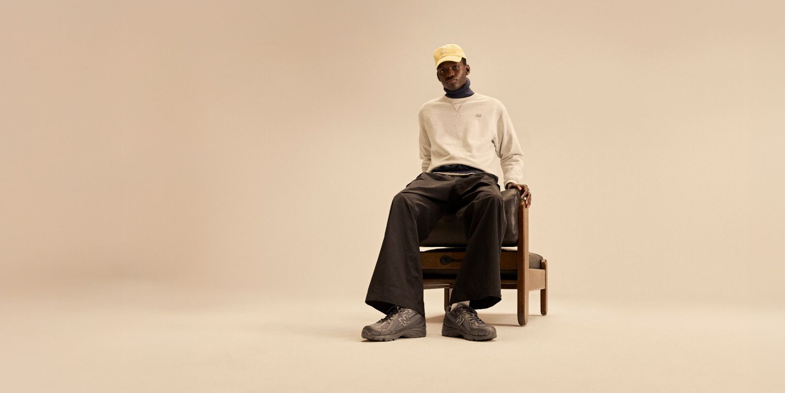 Model sitting on wooden and leather chair behind a beige background wearing a tan shirt, yellow hat, and dark New Balance 740s. 