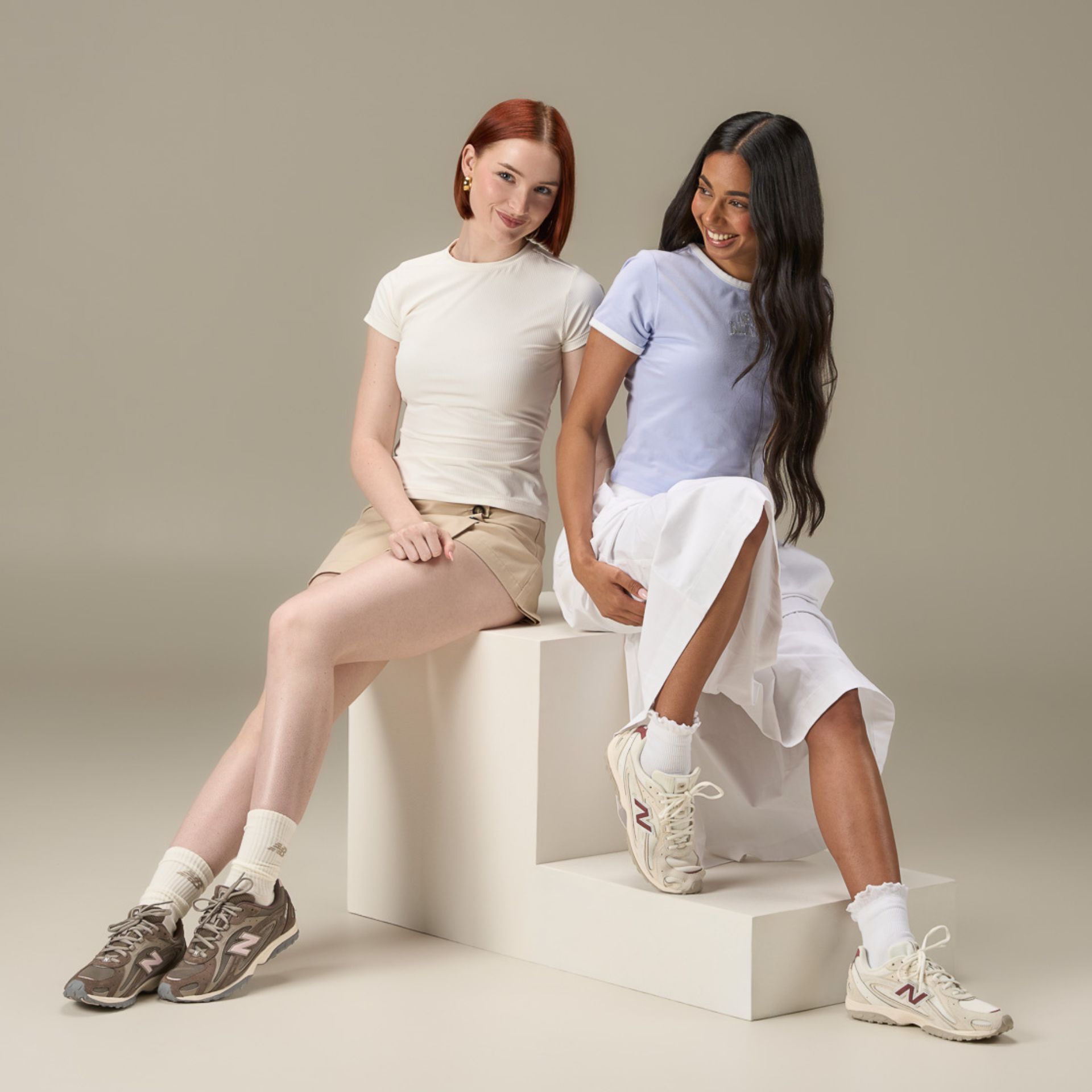 Two women sitting in a studio background wearing New Balance 204 sneakers
