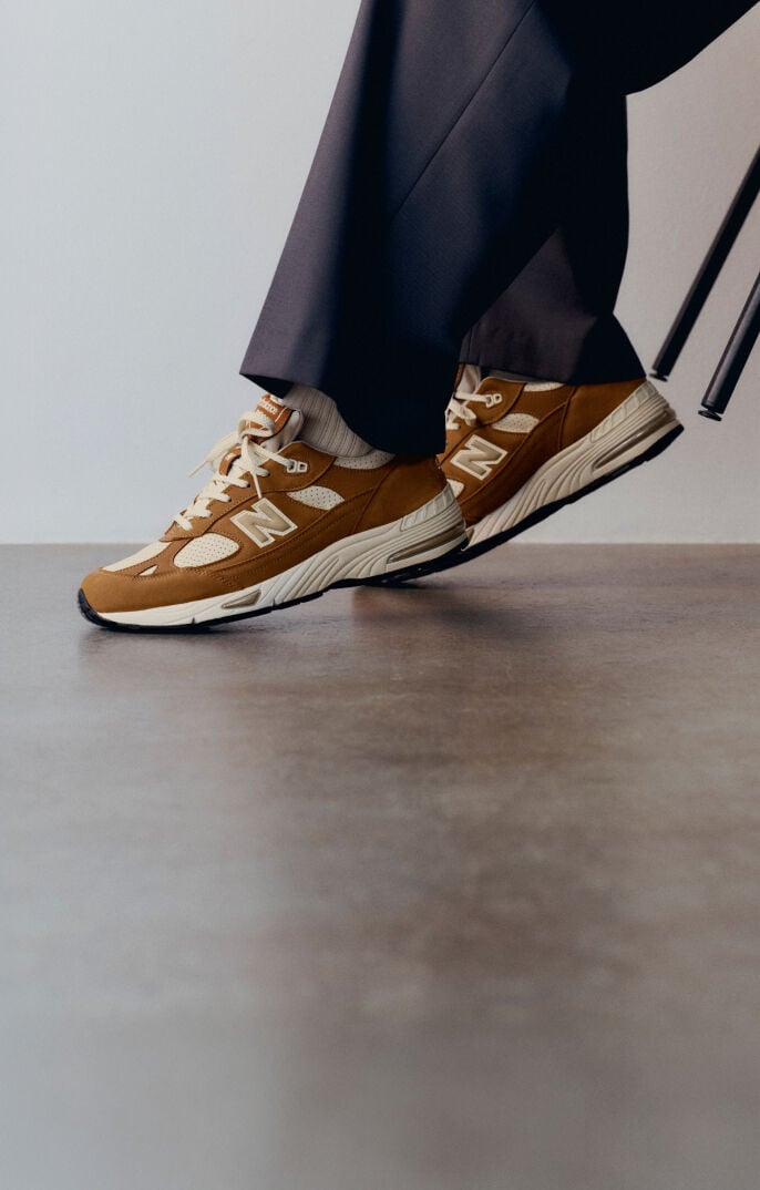 Knee down view of a woman wearing a pair of New Balance Made in UK sneakers in with room with a concrete floor and white walls. 